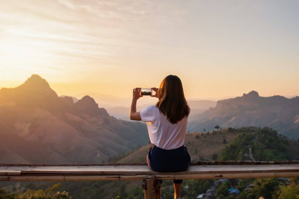 Young woman traveler taking photo with smart phone at sunset over the mountain